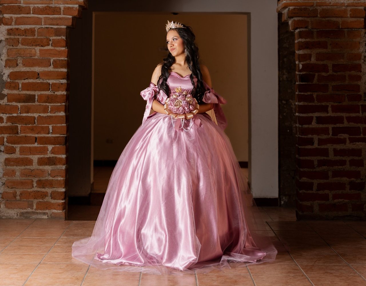 Quinceañera con vestido rosa y ramo de flores bajo un arco de ladrillo